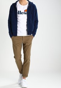 Marine blauwe zip-up hoodie, wit T-shirt met logo, khaki chino's, opgerolde mouwen, witte sneakers. Soepele textuur; casual pasvorm.