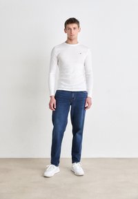 Vit långärmad skjorta, blå denimjeans och vita sneakers. Skjortan har en liten logotyp på bröstet. Enkelt mönster, slim fit.