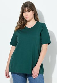 Tmavě zelené v-neck tričko vyrobené z jemné bavlny, s krátkým rukávem a volným střihem, v kombinaci s modrými džíny. Hladká textura s minimálními detaily.