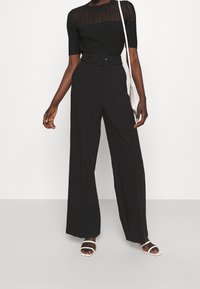Top corto a maniche corte nero a costine con una sezione superiore trasparente, abbinato a pantaloni neri a vita alta e gamba larga con una cintura coordinata.