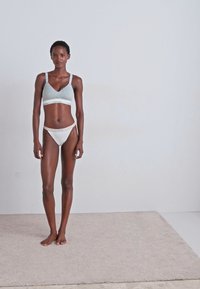 Világosszürke bralette vékony pántokkal és fehér elasztikus szalaggal, párosítva fehér tanga alsóneművel, a modell egy semleges árnyalatú texturált szőnyegen áll.