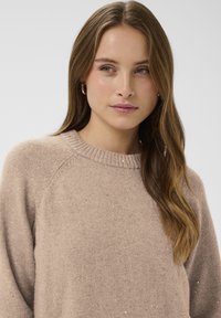 Bézs pulóver kerek nyakkivágással és raglán ujjakkal, puha textúrával és szórt, enyhe csillogó részletekkel.