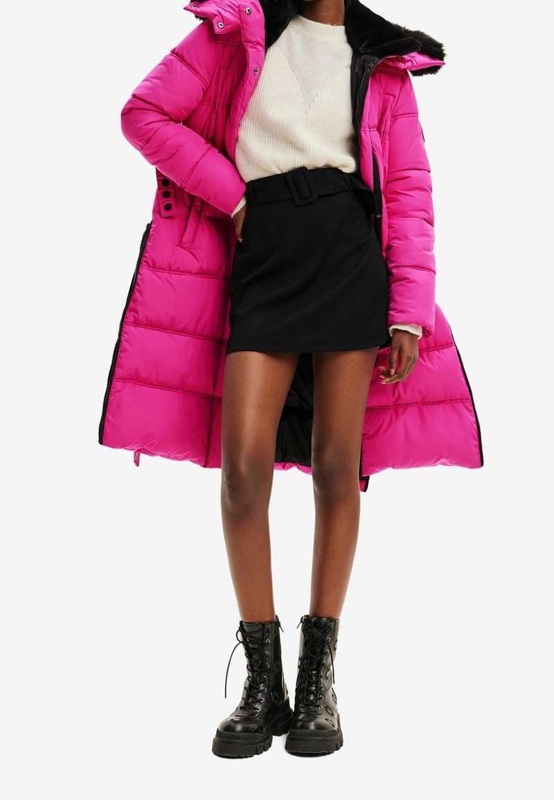 Femme portant un manteau matelassé rose vif par-dessus un pull blanc, une mini jupe noire avec ceinture, et des bottes de combat noires à lacets, debout devant un fond blanc.