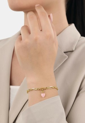Pulsera de cadena de oro con un charm en forma de corazón rosa. Enlaces gruesos con un acabado pulido, llevada en una muñeca sobre una camisa de color claro.