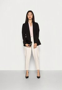Black blazer over a pink blouse, paired with light beige trousers. Holding a black clutch, wearing black heels. Straight, dark hair.