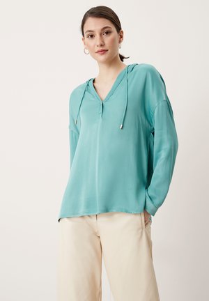 Jonge vrouw met bruin haar in een paardenstaart, gekleed in een teal lange mouwen hoodie-blouse en crèmekleurige wijde broek, tegen een effen achtergrond.