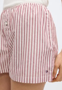 Spodenki w czerwono-kremowe paski, wykonane z lekkiego materiału, z elastycznym paskiem w talii, kieszeniami po bokach oraz małym logo na krawędzi.