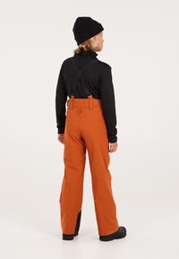 Pantaloni da sci arancioni con bretelle nere regolabili, vita alta e gambe larghe. Accenti neri alle caviglie. Indossati con una maglietta nera a maniche lunghe.