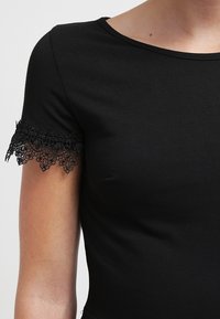 Top corto a maniche corte nero realizzato in un tessuto liscio. Presenta un delicato bordo in pizzo nero ai margini delle maniche, creando un contrasto testurizzato.