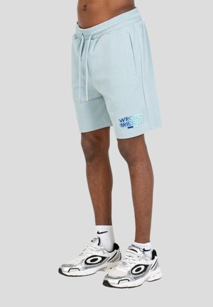 Uomo che indossa pantaloncini azzurri con la scritta "Wrong Friends" sulla gamba sinistra, calzini bianchi Nike e scarpe sportive bianche con dettagli neri.