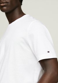 T-shirt bianco in cotone con scollo rotondo, maniche corte e un piccolo dettaglio del logo colorato sulla manica. Texture liscia e vestibilità classica.