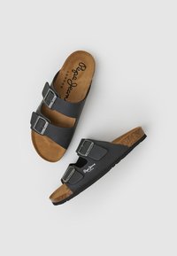 Crne klizne sandale s dva podesiva bucklea, podloga od pluta i mekani uložak sličan antilopu u smeđoj boji. Logo otisnut na donjoj traci.