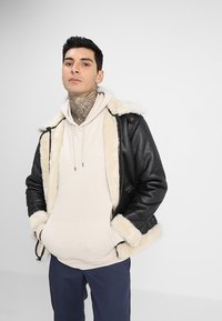 Zwarte leren jas met crèmekleurige neplederen voering en accenten, gedragen over een lichtbeige hoodie. Model draagt donkere broek.