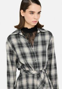 Ung kvinde med glat brunt hår iført en sort blondebluse under en sort-hvid ternet knapklædt kjole med et bundet bælte i taljen.