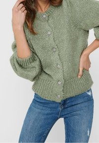 Zielony sweter z dzianiny z okrągłym dekoltem, teksturowany materiał, duże srebrne guziki i szerokie rękawy. Noszony na niebieskie zniszczone dżinsy.