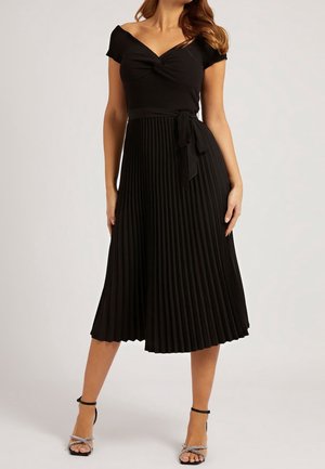 Frau trägt ein schwarzes schulterfreies Oberteil und einen plissierten Midirock mit gebundenem Taillengürtel, kombiniert mit schwarzen Riemchen-Sandalen mit hohen Absätzen.