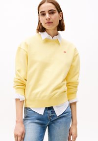 Geel crewneck sweatshirt van zacht materiaal, met een klein rood logo, gecombineerd met een wit overhemd eronder en blauwe jeans.