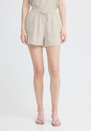 Femme portant un short beige à taille élastique avec un lien et des sandales tongs à motif peau de serpent, debout devant un fond blanc uni.