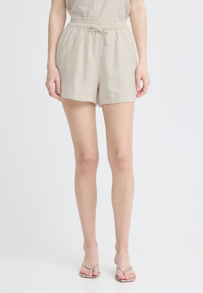 Femme portant un short beige à taille élastique avec un lien et des sandales tongs à motif peau de serpent, debout devant un fond blanc uni.
