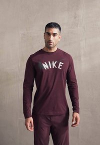 Vīrietis, tērpies bordo Nike garām piedurknēm krekliņā un atbilstošās biksēs, stāv pretī vienkāršai gaiši pelēkai teksturētai sieni.