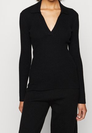 Kobieta w czarnym, prążkowanym, długim topie z głębokim dekoltem w serek i dopasowanych czarnych spodniach na jednolitym tle.