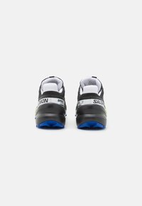 Schwarz-weiße Sportschuhe mit einer strukturierten Oberseite, Fersendämpfung in Schwarz und einer leuchtend blauen Laufsohle. Mit Logo und Branding.