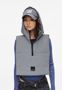 Gilet imbottito grigio con cappuccio, zip frontale e patch con logo nero. Indossato sopra un top a coste blu, abbinato a jeans scuri e a un cappello grigio.