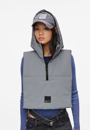 Grau gepolsterte Weste mit Kapuze, Reißverschluss vorne und schwarzem Logo-Patch. Getragen über einem blauen gerippten Oberteil, kombiniert mit dunklen Jeans und einer grauen Mütze.