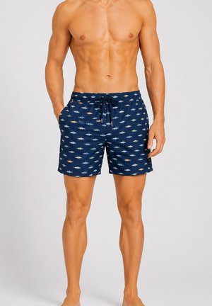 Homme musclé portant un short de bain bleu marine avec de petits motifs colorés de requins, debout les mains dans les poches devant un fond uni.