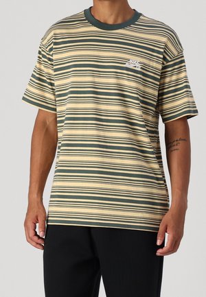 Mężczyzna w beżowo-ciemnozielonej pasiastym T-shircie Nike SB z krótkim rękawem i czarnych spodniach, widoczny tatuaż na prawej przedramieniu.