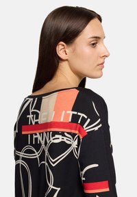 Czarny top z tkaniny z abstrakcyjnymi wzorami w kolorach beżowym, czerwonym i brzoskwiniowym, z szerokim dekoltem i kontrastowymi przeszyciami na ramionach.