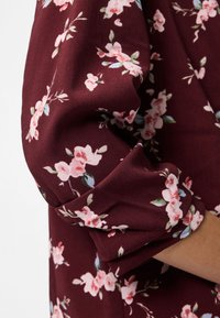 Erme av burgunderfarget stoff med rosa og hvitt blomsterpattern, lett opprullet, som viser deler av en persons arm.