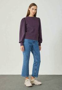 Lilla sviiter, millel on paisutatud varrukad ja soonikkoes allserv, koos siniste sirgete püksidega ja beežide spordijalatsitega, neutraalse taustaga.