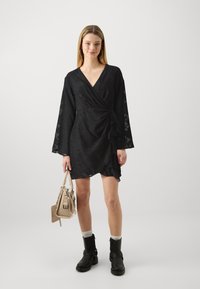 Μαύρο φορεμα με μακρύ μανίκι σε σχήμα καμπάνας, με υφή, και πλευρικό κόμπο. Συνδυασμένο με στρατιωτικά μποτάκια και μικρή μπεζ τσάντα.