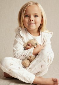 Langærmede, cremefarvede pyjamas med blomsterprint og flæse-detaljer, sammen med en blød, lysebrun bamse.