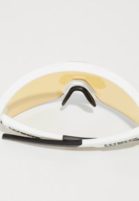 Sonnenbrille mit einem weißen Rahmen und gold getönten Gläsern. Verfügt über ein schwarzes Gumminasenpolster und schwarze Akzente an den Bügeln.