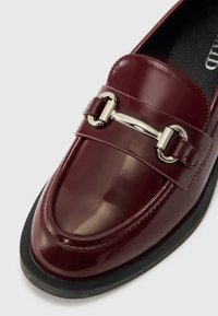 Burgundia läikiv nahk loafer, millel on ümar nina, hõbedase tooni riistvara klamber ja tekstureeritud must tald.