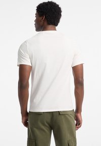 Ecrufarbenes T-Shirt mit kurzen Ärmeln und klassischem Rundhalsausschnitt, das eine lockere Passform bietet. Getragen mit olivgrünen Cargo-Hosen.