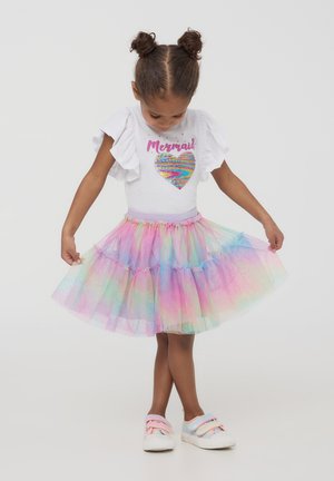 Top bianco con maniche a volant e un cuore di paillettes arcobaleno, abbinato a una gonna in tulle multicolore e sneakers dai colori pastello.