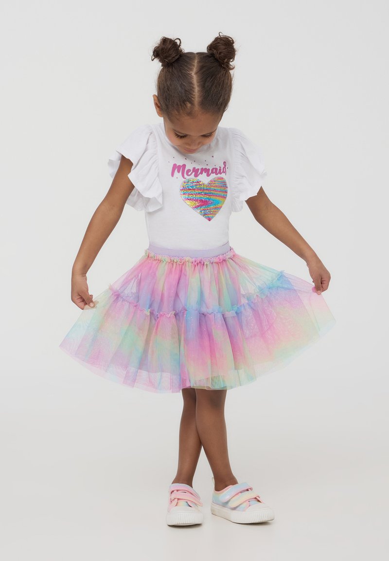 Top bianco con maniche a volant e un cuore di paillettes arcobaleno, abbinato a una gonna in tulle multicolore e sneakers dai colori pastello.