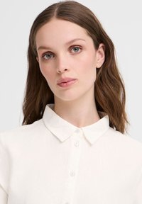 Camicia bianca con colletto, realizzata in tessuto leggero. Presenta una pattina dei bottoni parzialmente visibile e una texture liscia.