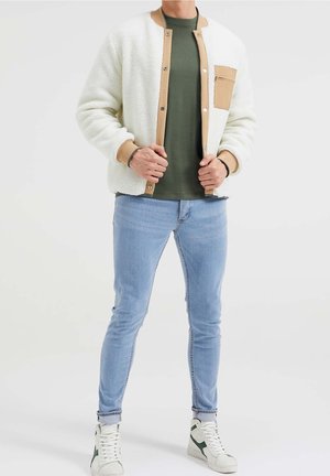 Giacca bomber in pile bianca con accenti beige, dotata di chiusura a bottoni e tasca sul petto. Abbinata a jeans skinny azzurri e sneaker bianche.