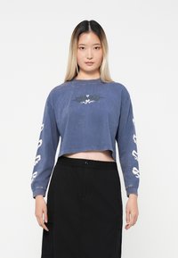 Skrajšan dolg rokav majica iz bledomodrega bombaža, ki ima črn grafični dizajn na prsih in bele napise na obeh rokavih.