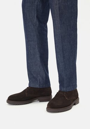 Brązowe zamszowe botki za kostkę z zaokrąglonym noskiem, sznurowadłami i gumowymi podeszwami, zestawione z ciemnymi denimowymi spodniami o delikatnej fakturze.