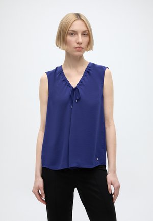 Femme aux cheveux blonds courts portant un chemisier bleu roi sans manches avec un détail noué à l'encolure et un pantalon noir, debout sur fond blanc.