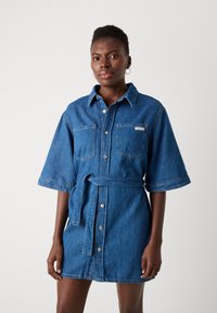 Denim srajčna obleka s kratkimi rokavi, pasom, dvema žepoma na prsih in srebrnimi gumbi, v srednji modri pranju. Gladka textura.