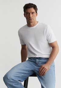 Mand iført en ensfarvet hvid kortærmet T-shirt og lyseblå jeans, der sidder på en sort skammel foran en hvid baggrund.