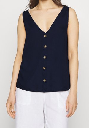 Frau trägt ärmelloses, marineblaues V-Ausschnitt-Oberteil mit fünf Knöpfen vorne und weiße Hose mit großen Taschen.