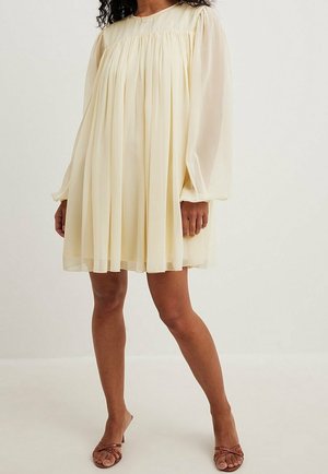 Mujer con un vestido mini holgado de manga larga de color amarillo pálido con tela fruncida y sandalias marrones de tacón con tiras, de pie frente a un fondo liso.