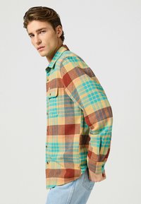 Camisa de franela multicolor con patrones de cuadros en amarillo, azul y rojo. Cierre de botones y un bolsillo en el pecho. Corte casual, mangas largas.